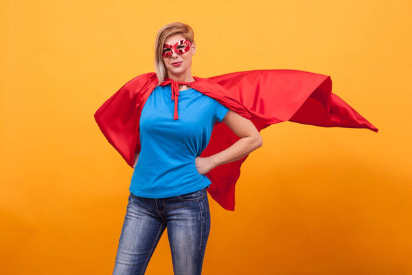 Young woman in superheros costume standing proudly with her red cape in the air over yellow background