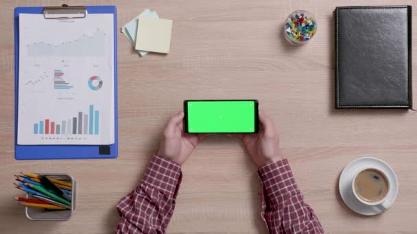 Manos de hombre sosteniendo verticalmente un teléfono inteligente con pantalla verde sobre un escritorio de oficina — Vídeos de Stock