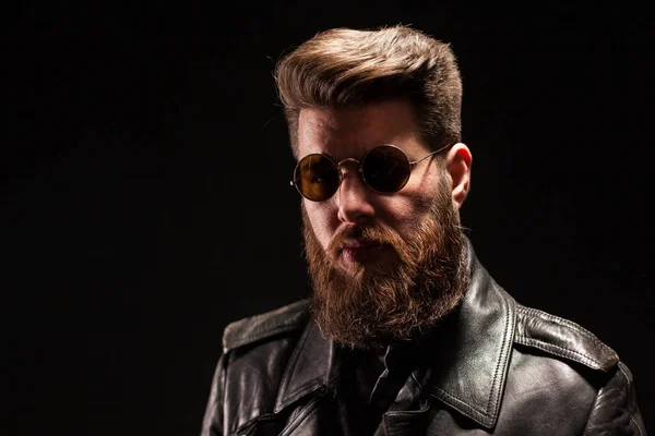 Homme barbu élégant avec veste en cuir et lunettes de soleil sur fond noir — Photo