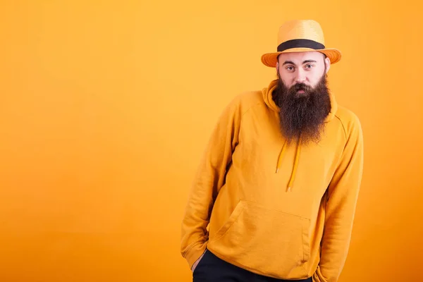 Cool bearded hipster with orange hat holding hands in his pockets over yellow background — Stock Photo, Image