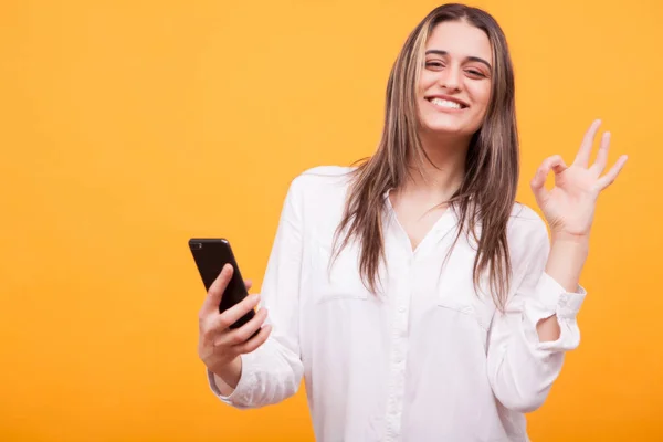 Mooi meisje in wit shirt houden telefoon en toont OK teken over gele achtergrond — Stockfoto
