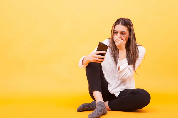Meisje op zoek geschokt op haar telefoon terwijl zitten over gele achtergrond — Stockfoto