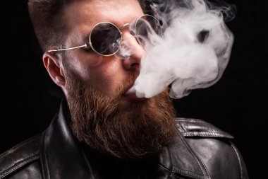 Siyah deri ceket giyen ve siyah arka plan üzerinde elektronik sigara içen sunglases emin sakallı adamın portresi kadar yakın