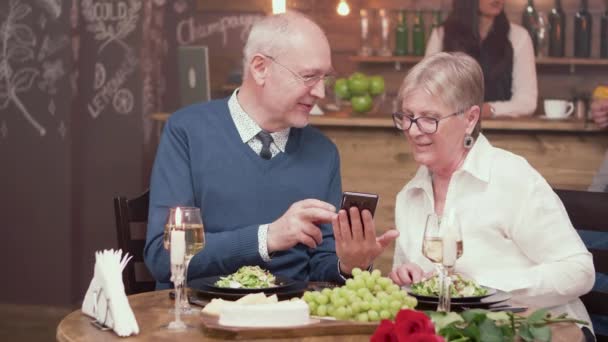 Gorgeous old couple on a date in vintage restaurant looking through photos on a smartphone — Stock Video