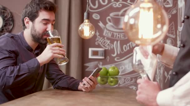 Молодой красивый мужчина за барной стойкой пьет пиво и смотрит на свой смартфон — стоковое видео