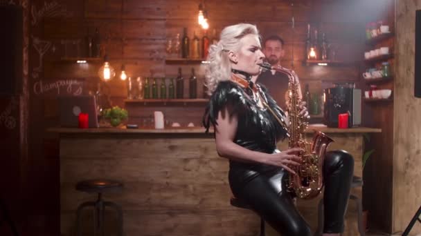 Young blonde female musician performs a song on her saxophone sitting on a high bar chair — 비디오