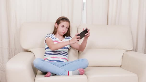 Chica adolescente en un sofá jugando un juego en un teléfono inteligente — Vídeos de Stock