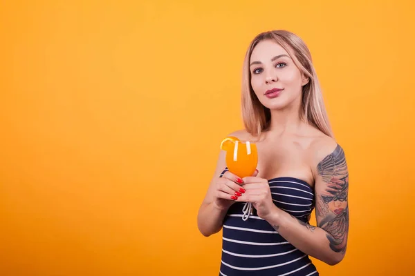 Сексуальна молода біла жінка в купальнику тримає склянку свіжого коктейлю і дивиться на камеру на жовтому тлі — стокове фото