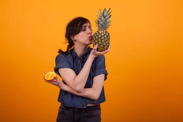 Retrato de una guapa jovencita besando ananas y sosteniendo una naranja fresca en el estudio sobre fondo amarillo — Foto de Stock