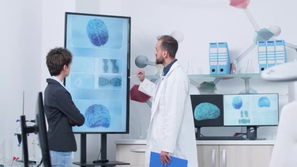 Doctor in white robe in medical research facility showing a 3D brain simulation to a patient — Stock Video