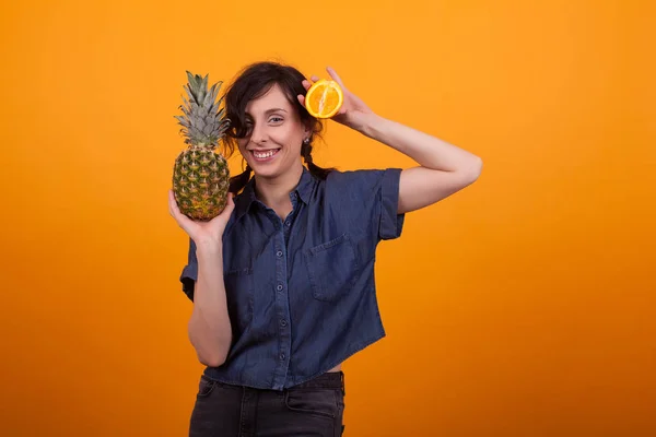 Atractiva joven mujer sonriendo a la cámara y sosteniendo frutas exóticas en el estudio sobre fondo amarillo — Foto de Stock