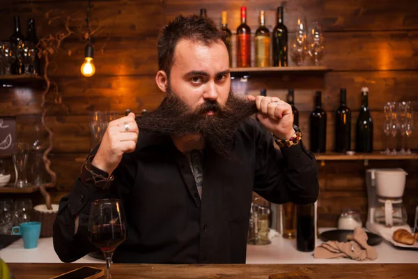 Barman atraente jogando com sua longa barba atrás do balcão . — Fotografia de Stock