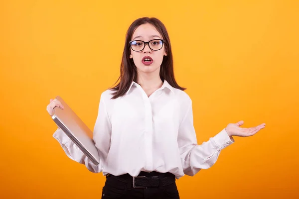 Ritratto di giovane ragazza carina con gli occhiali cercando confuso e tenendo il computer portatile in studio su sfondo giallo — Foto Stock