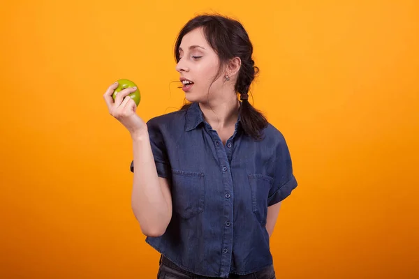 Sido porträtt av glad ung kvinna som håller och tittar på ett grönt äpple i studion över gul bakgrund — Stockfoto