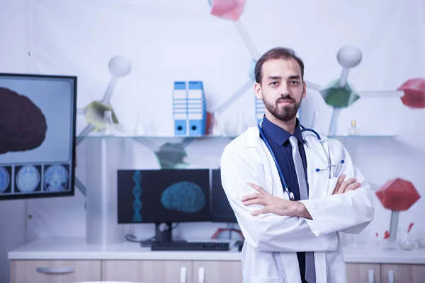 Atractivo médico barbudo con los brazos cruzados mirando a la cámara en su gabinete del hospital —  Fotos de Stock