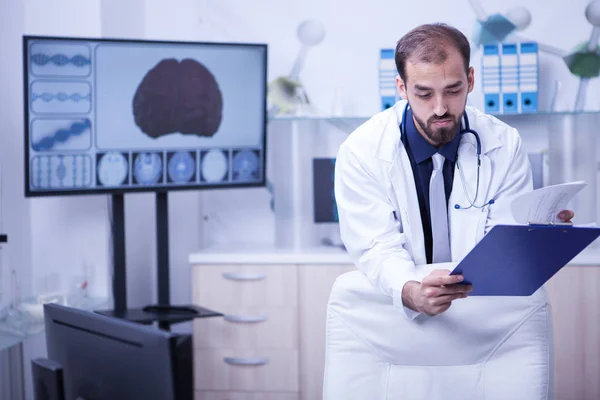 Hombre doctor en su gabinete desde el hospital mirando sus notas desde el portapapeles — Foto de Stock