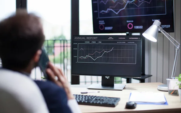 Young marketing expert kijkt naar grafics op de monitor en heeft een zakelijk gesprek op zijn telefoon — Stockfoto