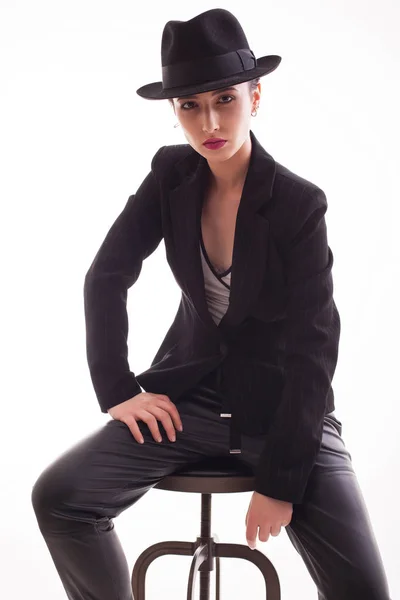 Modèle de mode élégant posant assis sur une chaise portant un chapeau rétro sur un fond blanc en studio — Photo