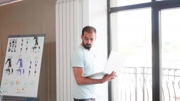 Kreativní muž, který pracuje na přenosném počítači vedle okna a pohybuje se ve své kanceláři — Stock video
