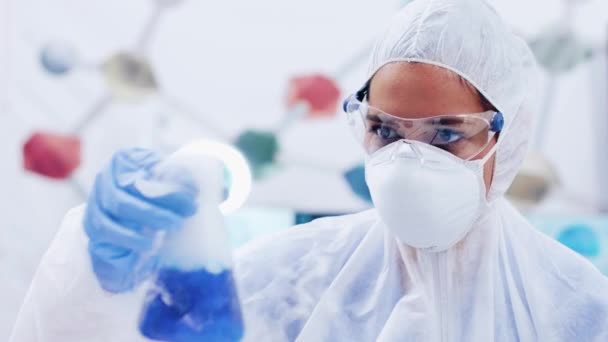Γυναίκα επιστήμονας σε προστατευτικό εξοπλισμό κουνώντας ένα μπουκάλι με μπλε υγρό καπνίσματος — Αρχείο Βίντεο