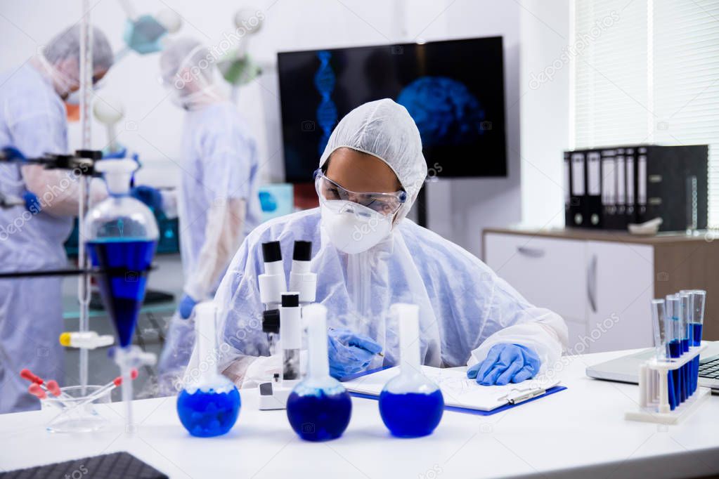 Female scientist in protection equipment taking notes with smoking blue solution