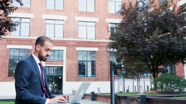 Бизнесмен на открытом воздухе печатает на ноутбуке, сидя в деловом районе — стоковое видео