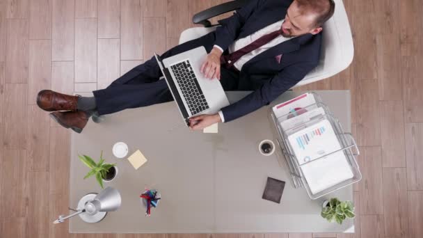 Вид зверху молодого бізнесмена в костюмі, який тримає ноги на своєму офісному столі — стокове відео