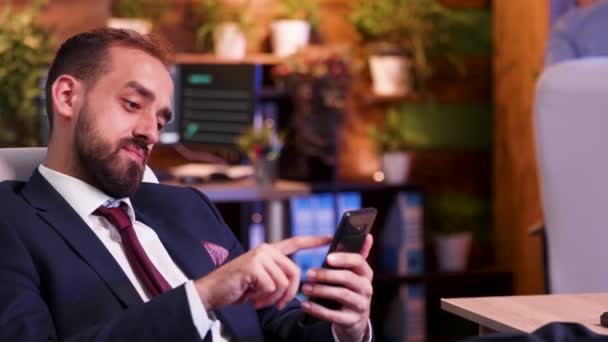Homem de negócios relaxado no escritório à noite rolar para baixo em seu telefone — Vídeo de Stock