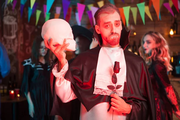 Gruseliger Mann im Dracula-Kostüm mit Totenkopf in der Hand, der bei der Halloween-Party in die Kamera schaut — Stockfoto