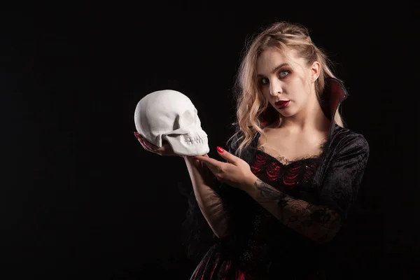 頭蓋骨を保持し、暗い背景に隔離されたハロウィーンのためのカメラを見てセクシーな吸血鬼の女性 — ストック写真