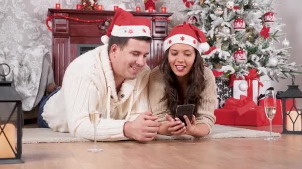 Pareja sonriente sentada en el piso de compras en línea usando un teléfono inteligente, habitación decorada con Navidad — Vídeos de Stock