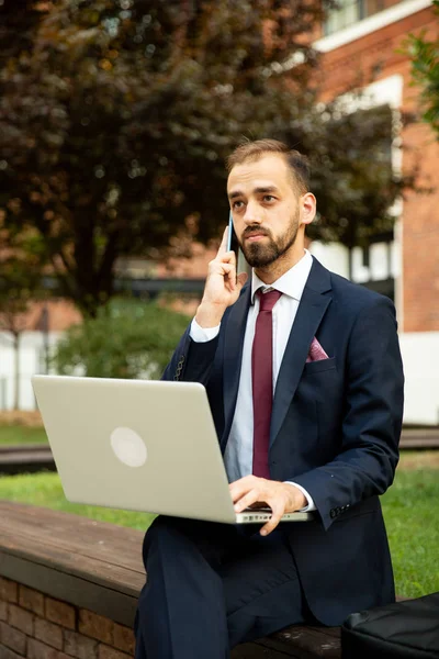 Jovem empresário trabalhando no laptop e falando ao telefone em um espaço verde — Fotografia de Stock