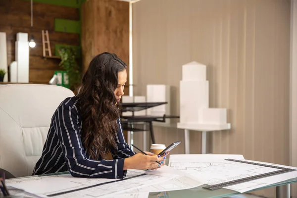 Architektin sitzt am Tisch, hält Smartphone in der Hand und tippt etwas drauf — Stockfoto