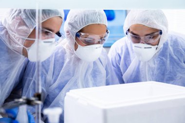 Üç bilim adamı laboratuvarda beyaz bir kutuya bakıyor.