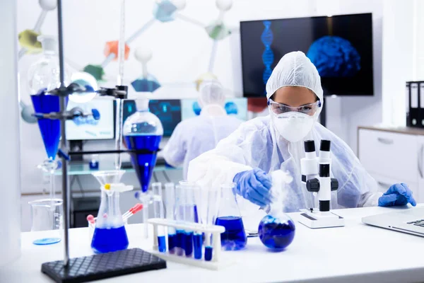 Cientista feminina com um microscópio no fron de sua alcançando para fora para um tubo de teste com solução azul — Fotografia de Stock