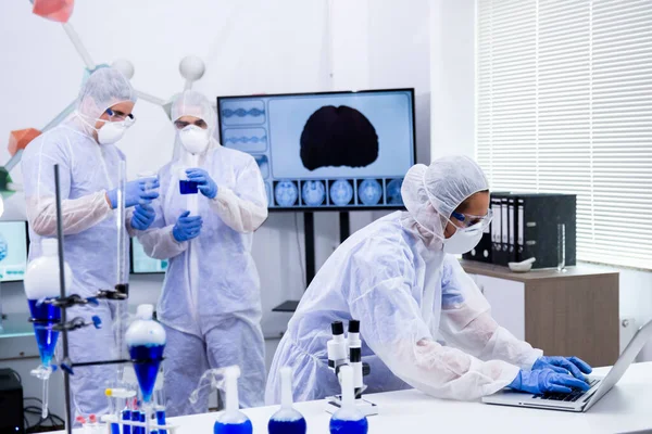 Женщина-ученый работает с ноутбуком и коллегами, работающими с реактивным голубым раствором — стоковое фото
