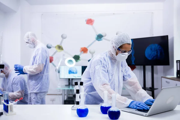 Científica femenina escribiendo en el portátil con solución azul humeante de tubos de ensayo en frente de su — Foto de Stock