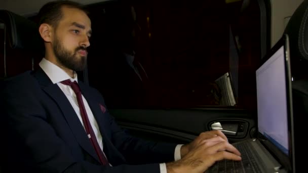Успешный бизнесмен печатает на своем ноутбуке на заднем сиденье своего лимузина — стоковое видео