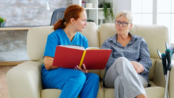 Caucasian female assistant in nursing home reading a book to an elderly retired woman — Stock Photo, Image