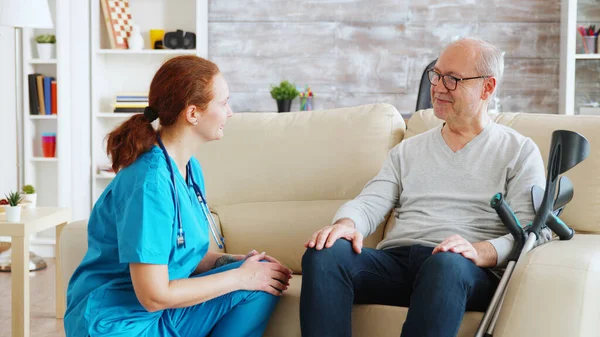 Female nurse talking and taking care of old disabled man sitting on the sofa — Stock Photo, Image