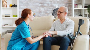 Bayan hemşire konuşuyor ve koltukta oturan engelli adamla ilgileniyor.