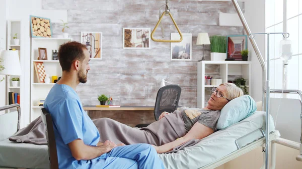 Male assistant taking a seat near hospital bed of ill lady lying in nursing home — Stock Photo, Image