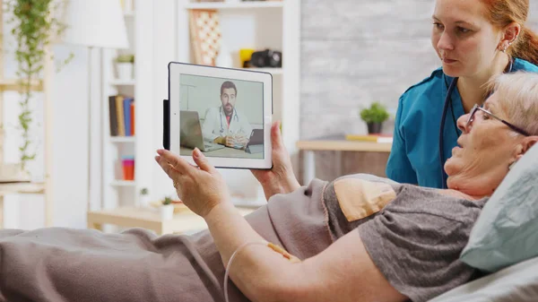 Old disabled woman lying in hospital bed having an online video call with a doctor — Stock Photo, Image