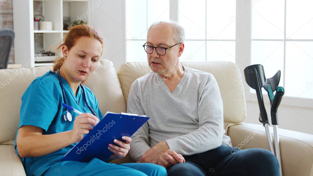 Female healthcare assistant taking notes about an old man health in cozy nursing home