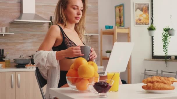 Сексуальная женщина в провокационном белье держит ноутбук, наслаждаясь кофе по утрам — стоковое видео