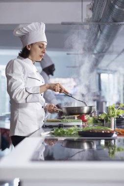 Mutfak mükemmeliyetine odaklanmış, üniformalı kadın şef ve şapkalı aşçı ocağındaki buharlı tencerede malzemeleri karıştırıyor, yemek endüstrisi için hassas ve tutkulu bir gurme tabağı hazırlıyor..