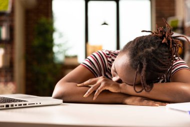 Laptopta çok çalıştıktan sonra masa başında uyuyan Afrikalı Amerikalı bayan girişimci. Yorgun siyah iş kadını, evde geçirdiği uzun saatlerden sonra dinlenmek için kollarının üzerinde dinleniyor..