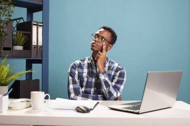 Elleri yanağında Afrika kökenli Amerikalı bir yönetici, yukarı bakıyor ve derin düşünceler içinde, ofis raporlarını yansıtıyor. Siyah adam masada oturuyor, yüzüne dokunuyor ve iş fikirlerini düşünüyor..