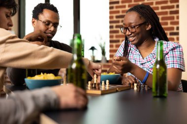 Afro-Amerikalı kadın ve erkek gülümseyerek arkadaşlarının satranç taşını masaya koymasını izliyorlar. Çok ırklı erkek ve kadın arkadaşlar içki ve atıştırmalıklarla oturdular ve gündelik masa oyunlarının keyfini çıkardılar..