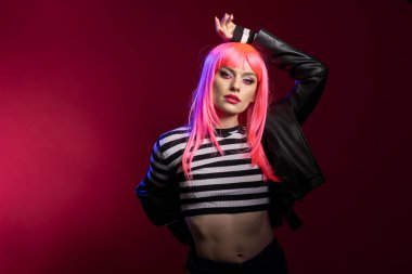 Renkli saçlı ve deri ceketli çekici bir kadın model modern stüdyoda tek koluyla poz veriyor. Çizgili tişörtlü şık, beyaz kadın kameraya doğrudan bakıyor..